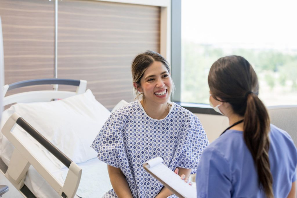 The young adult woman sits up in her hospital bed with a big smile on her face when she gets the good test results from the unrecognizable female nurse.
