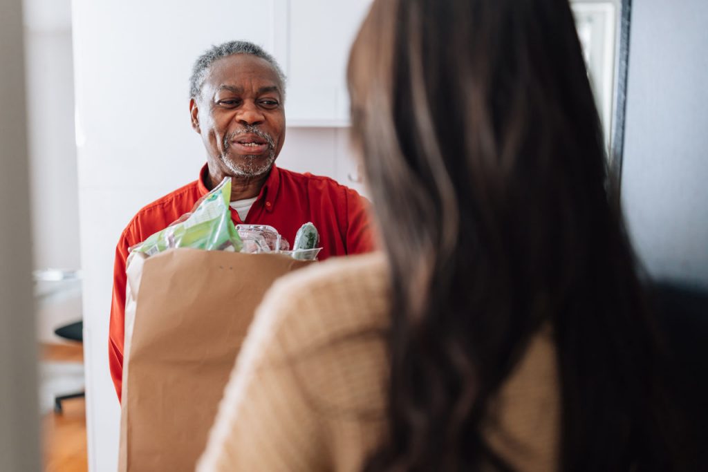 A young female Caucasian volunteer brings bag with food to an older man's front door