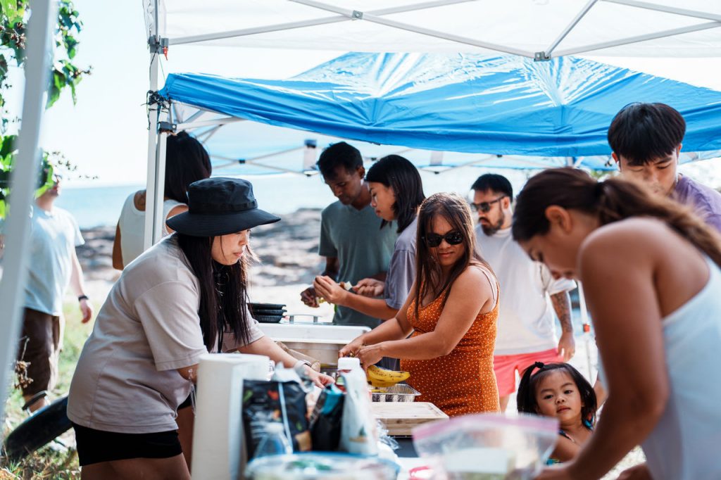A multiracial group of adults and kids gather around tables of food while having a barbecue lunch at the beach in Hawaii.