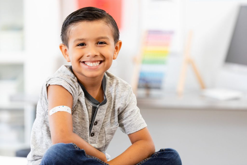 A cheerful boy with a bandage on his arm smiles broadly, showing his toothy smile.  He looks happy and healthy.