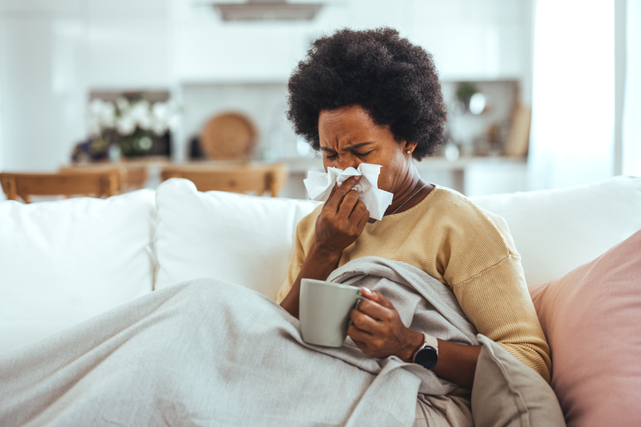 Sick young woman sitting on sofa blowing her nose at home in the sitting room. Photo of sneezing woman in paper tissue. Picture showing woman sneezing on tissue on couch in the living-room